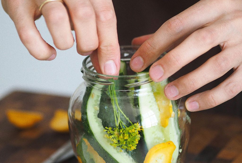 The photo shows people putting cucumber and spices in a glass jar.