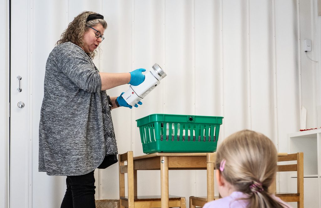 Henkilö käsittelee käsineet kädessä kädessä ilmanäytelaitetta, ja lapsi istuu etualalla seuraamassa tilannetta sisätilassa.