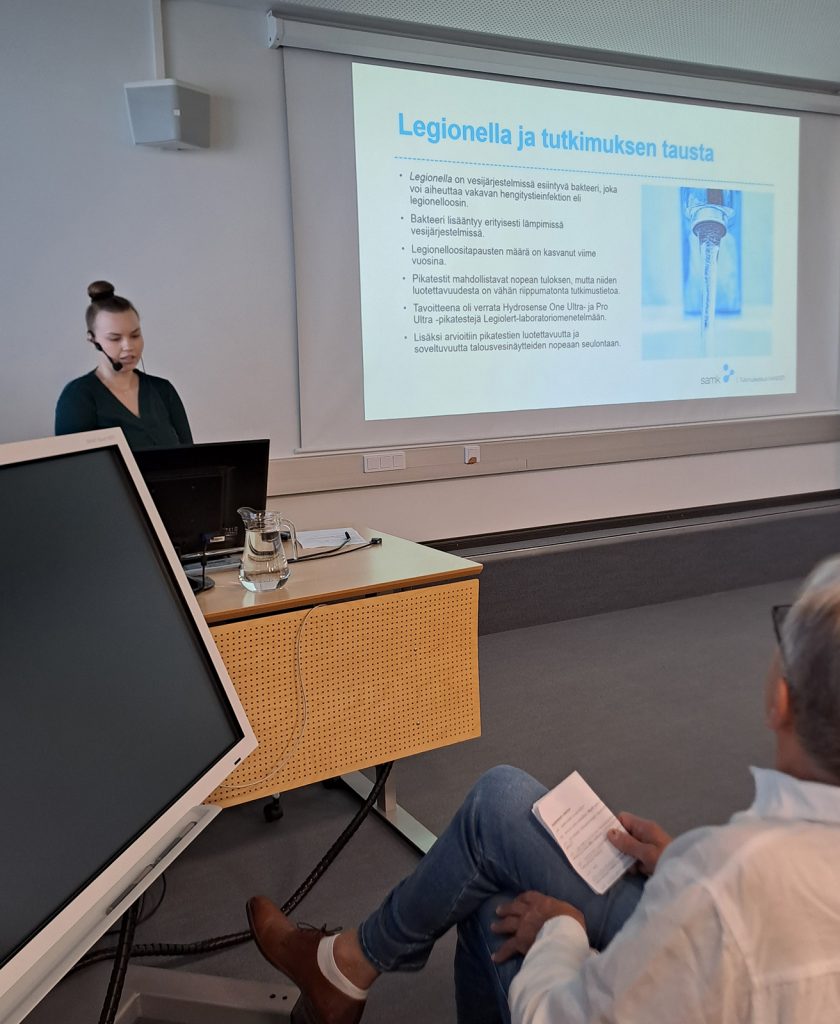 Henkilö pitää esitystä luentosalissa legionellabakteereita käsittelevän diasarjan äärellä.