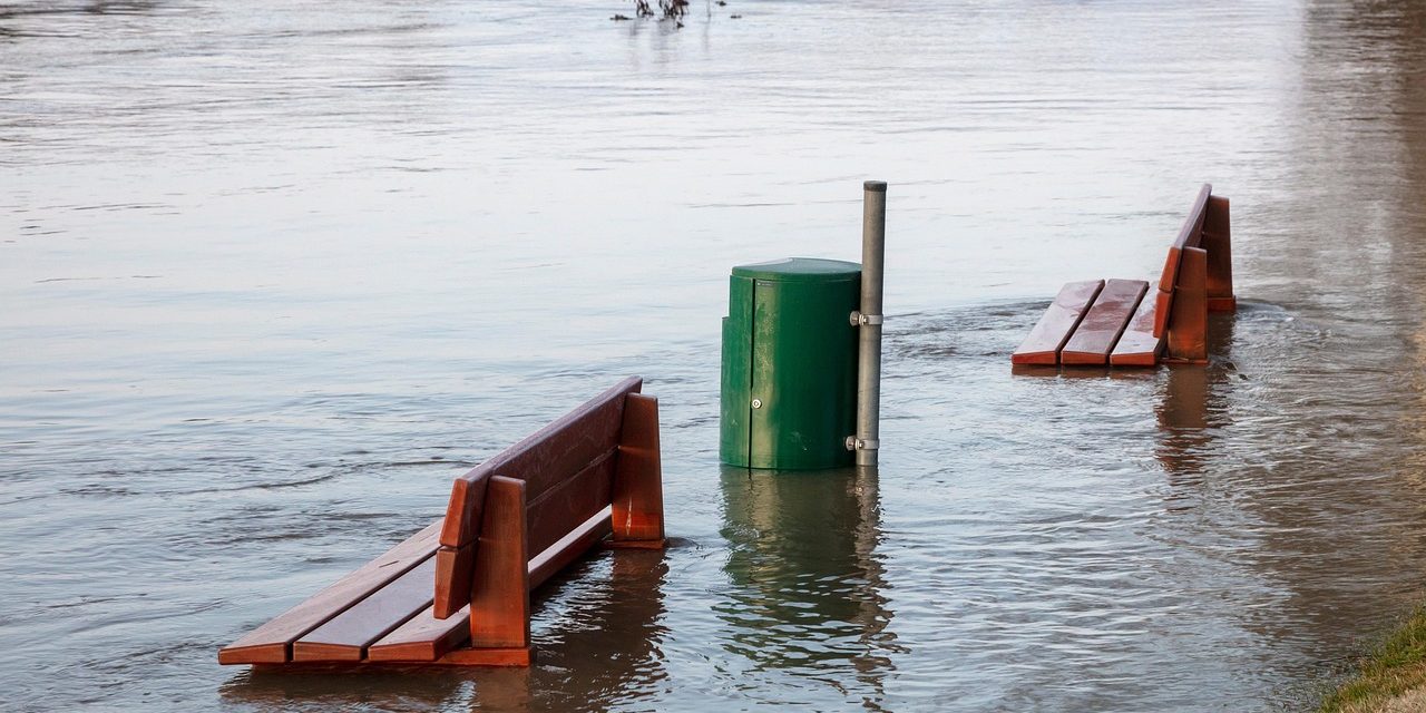 In the picture, two benches and a trash can are half covered by floodwater.